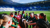 A group of football players in green jerseys huddled together on a stadium field with empty stands in the background.