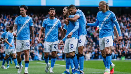 Manchester City players celebrating a goal wearing the team's blue jerseys with Etihad Airways sponsorship at a crowded stadium.