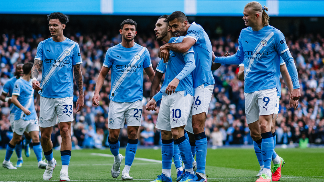 Manchester City players celebrating a goal wearing the team's blue jerseys with Etihad Airways sponsorship at a crowded stadium.