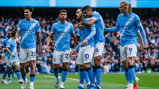 Manchester City players celebrating a goal wearing the team's blue jerseys with Etihad Airways sponsorship at a crowded stadium.