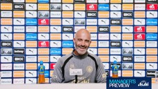A Manchester City coach smiling during a press conference with a backdrop of various sponsor logos and branding.