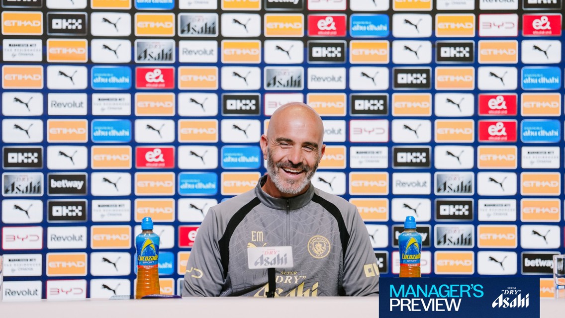 A Manchester City coach smiling during a press conference with a backdrop of various sponsor logos and branding.