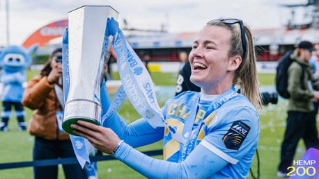 A smiling football player in a blue jersey holds a trophy with Barclays ribbons on a football field. Mascots and photographers are in the background.
