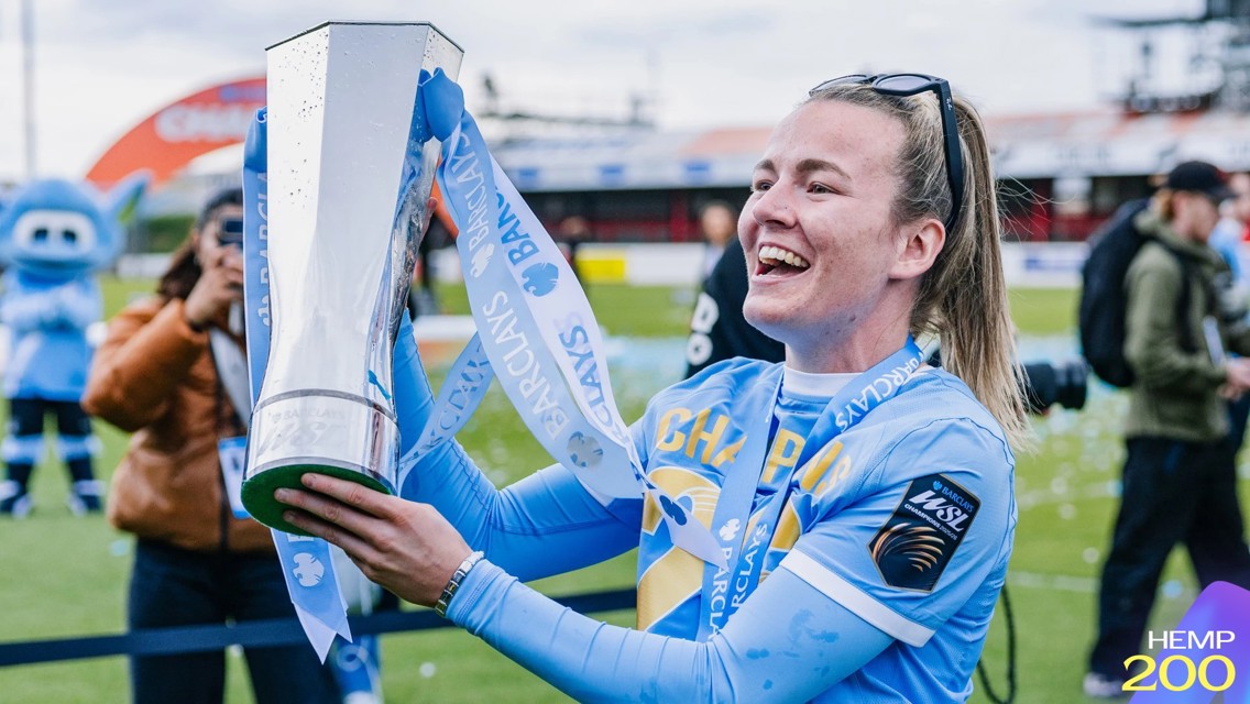 A smiling football player in a blue jersey holds a trophy with Barclays ribbons on a football field. Mascots and photographers are in the background.