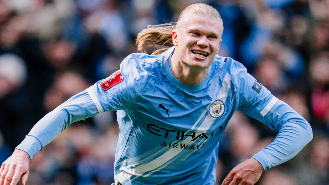 A footballer in a blue Manchester City jersey in action during a match, blurred crowd in the background.