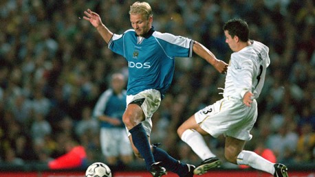 Two football players in action during a match, one in a blue Manchester City jersey and the other in a white Leeds United jersey, near a football.