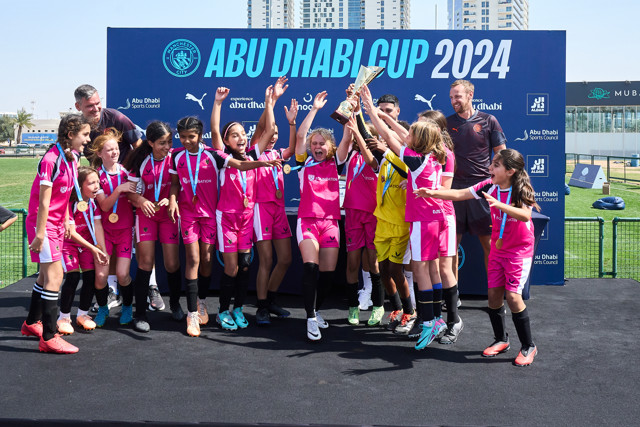 A girls' soccer team celebrates winning the Abu Dhabi Cup 2024, holding a trophy and wearing medals. The players are dressed in pink uniforms.