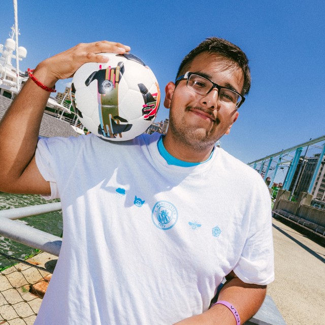 A person holding a soccer ball near their shoulder, standing beside a waterfront with a large ship in the background. The person is wearing glasses and a white t-shirt with blue logos.