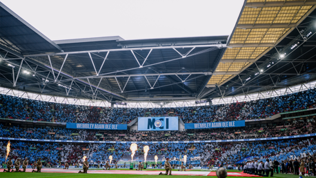 Wide view of Wembley Stadium with fireworks and a crowd. Banners with the Manchester City crest are visible.