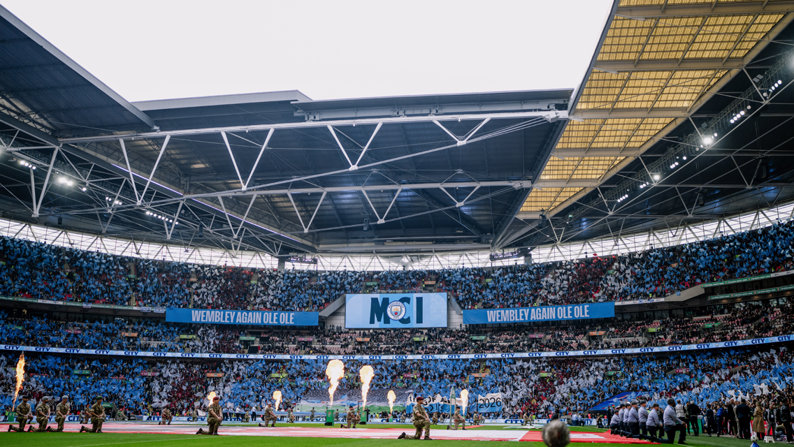 Wide view of Wembley Stadium with fireworks and a crowd. Banners with the Manchester City crest are visible.