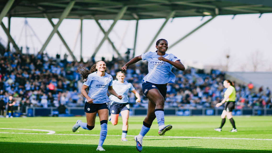 Two Manchester City women's players celebrating on the field after a goal with a crowd in the background.