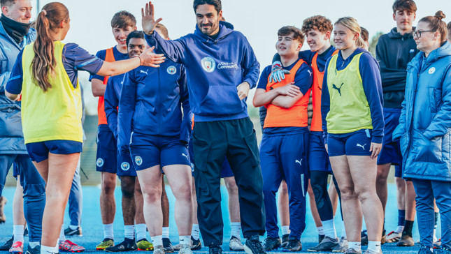 A group of Manchester City players and a coach engaging in a high five during a training session. Players are wearing team apparel including Puma kits and 'City in the Community' hoodies.