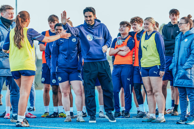 A group of Manchester City players and a coach engaging in a high five during a training session. Players are wearing team apparel including Puma kits and 'City in the Community' hoodies.
