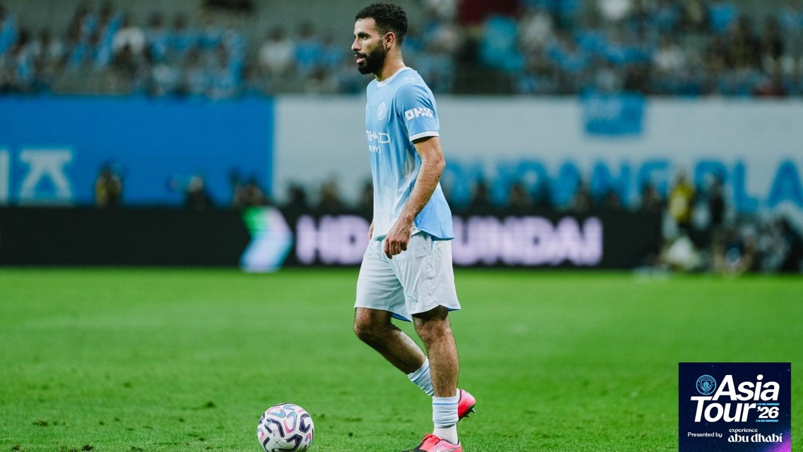 A soccer player in light blue kit stands on a field with a ball during the Asia Tour '26 event. The background shows blurred spectators and advertising boards.