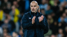 A bald man wearing a dark jacket is clapping, with a crowd blurred in the background.