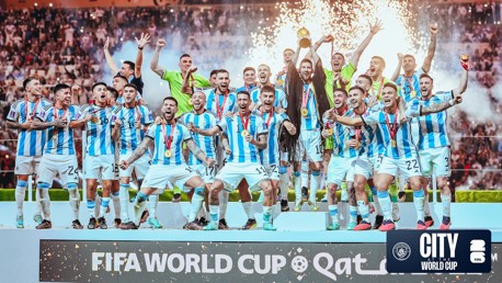 A football team in blue and white stripes celebrating with the World Cup trophy on a platform, surrounded by fireworks and a cheering crowd. The FIFA World Cup and Qatar text are visible.