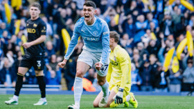 Manchester City player celebrates scoring a goal during a match. The background shows a crowd cheering and another team's goalkeeper kneeling on the ground.