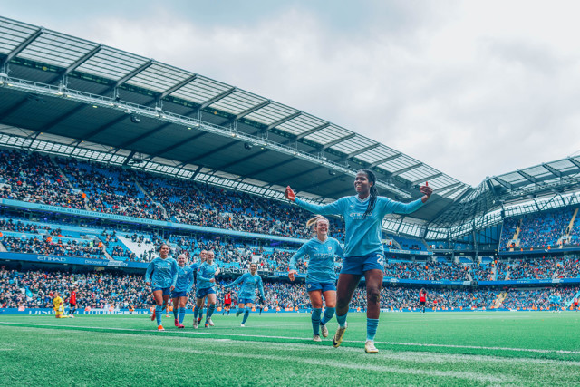 Manchester City women's team players celebrating at Etihad Stadium during a match, with a large crowd in the background.