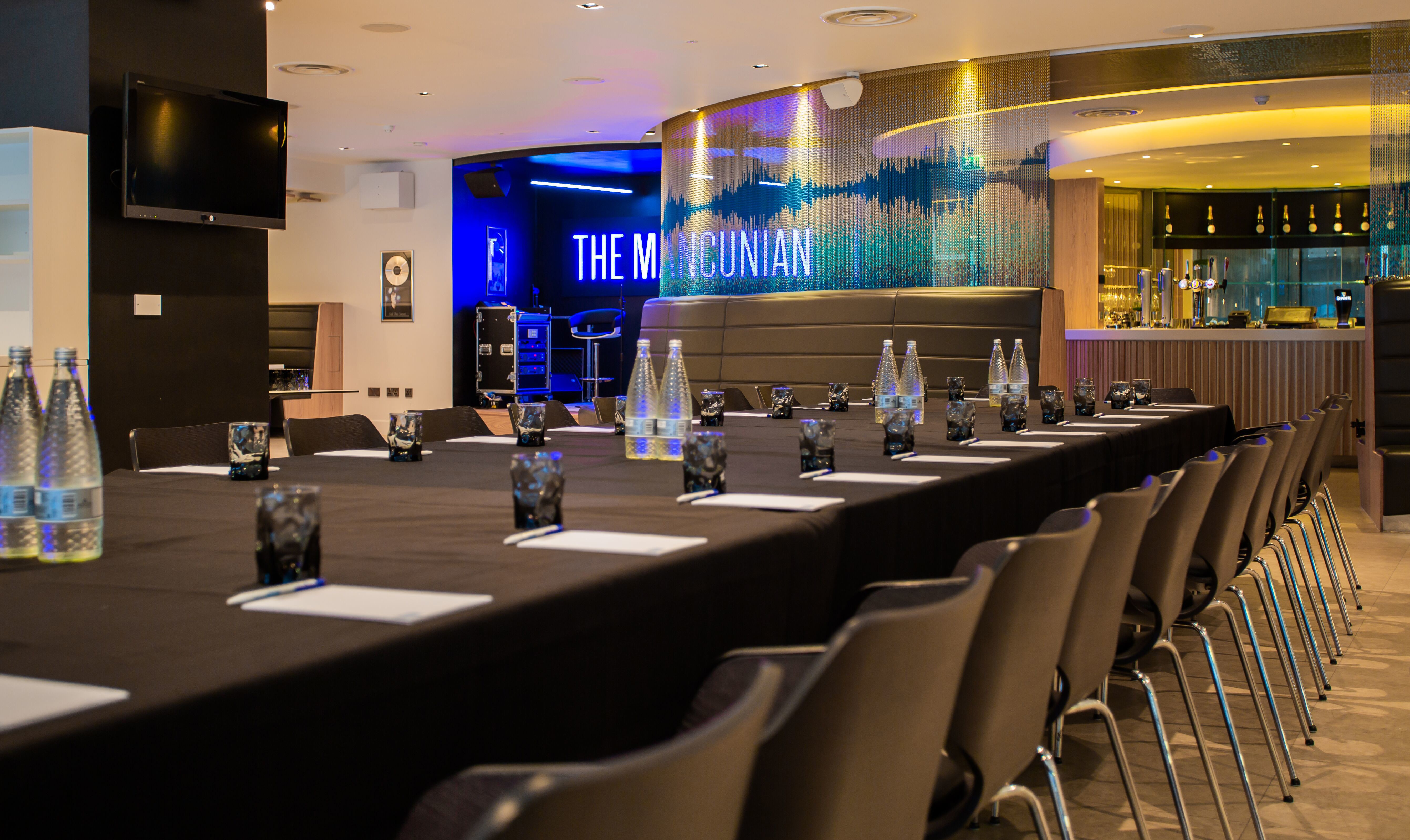 A conference room setup with long tables, chairs, and water bottles. The wall has 'THE MANCUNIAN' written on it, and there is a bar area in the background.