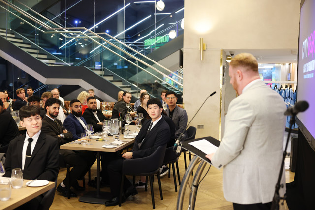 A speaker addresses an audience in a formal dining setting with people seated in suits listening attentively.