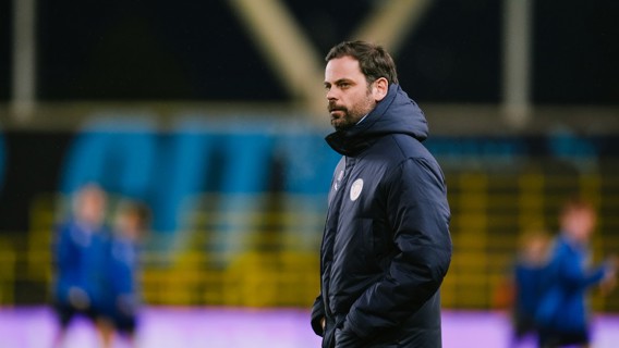 A man wearing a Manchester City jacket observes a training session on a sports field. The background shows players in motion, likely during a warm-up routine.