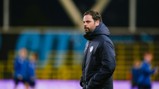 A man wearing a Manchester City jacket observes a training session on a sports field. The background shows players in motion, likely during a warm-up routine.