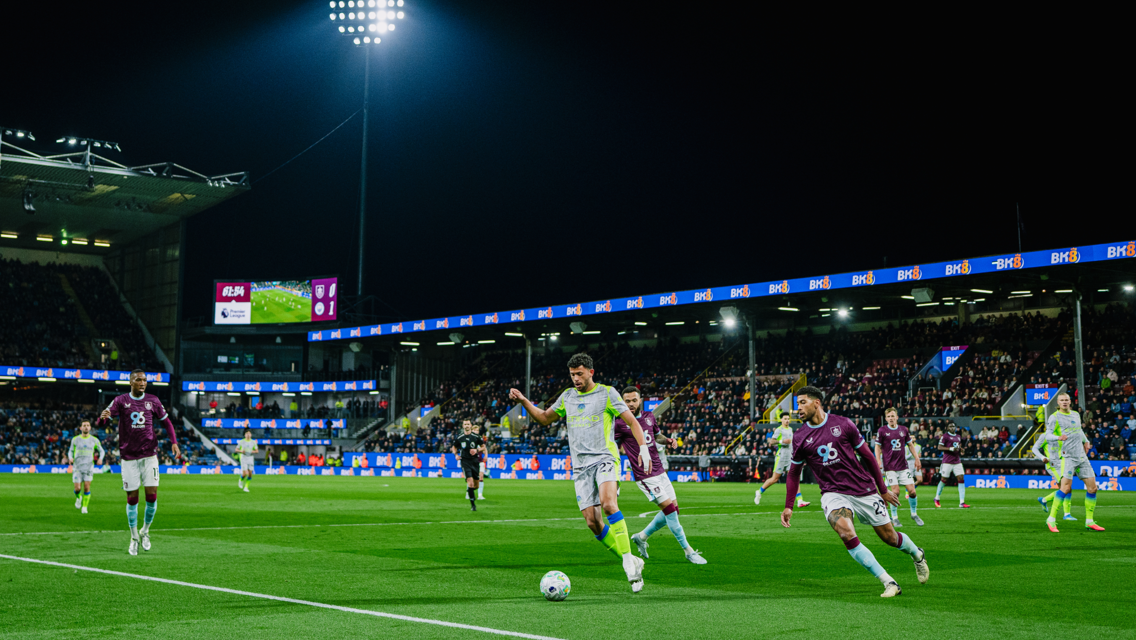 Soccer players from two teams competing under stadium lights, one team in claret jerseys and the other in light-colored kits.