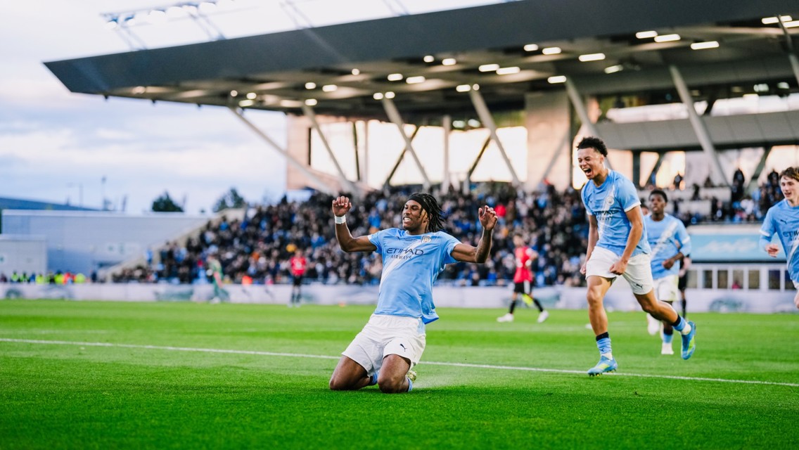 A football player wearing a light blue jersey kneels on the field, celebrating a goal with arms raised. Teammates run towards him with excitement. The stadium is filled with spectators.