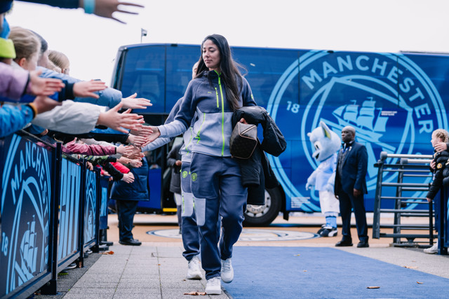 A Manchester City player arrives for matchday, walking past and high-fiving fans. The Manchester City team bus and mascot are visible in the background.
