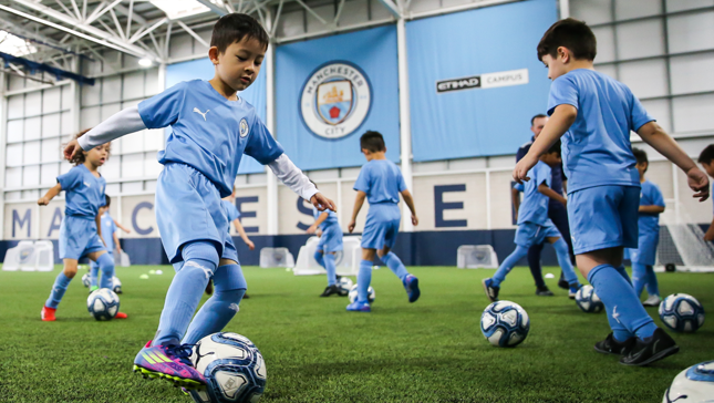 Young players in Manchester City kits training indoors on an artificial turf field with multiple soccer balls.