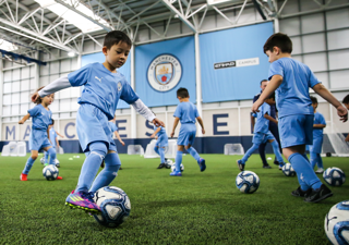 Young players in Manchester City kits training indoors on an artificial turf field with multiple soccer balls.