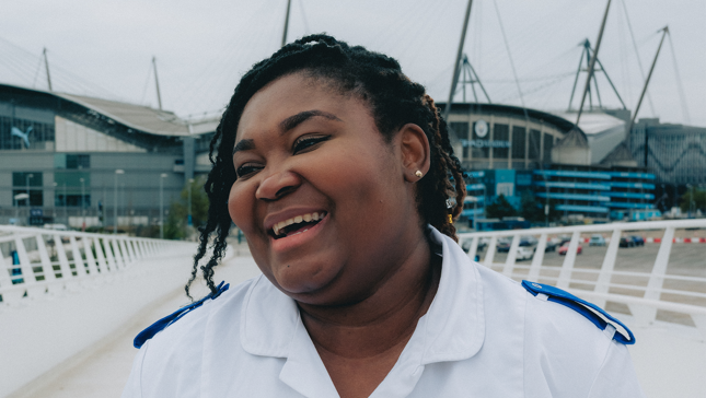 A person in a white outfit smiling in front of the Etihad Stadium, home to Manchester City.