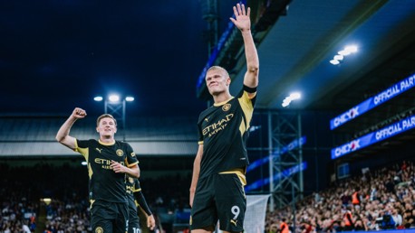 Two Manchester City players celebrating a goal on the field, one raising his hand and the other with a fist in the air. The stadium is filled with spectators.
