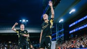 Two Manchester City players celebrating a goal on the field, one raising his hand and the other with a fist in the air. The stadium is filled with spectators.