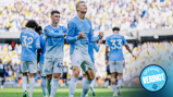 A group of Manchester City players celebrating a goal on the field. One player is prominently fist-pumping while others in light blue kits are in the background.