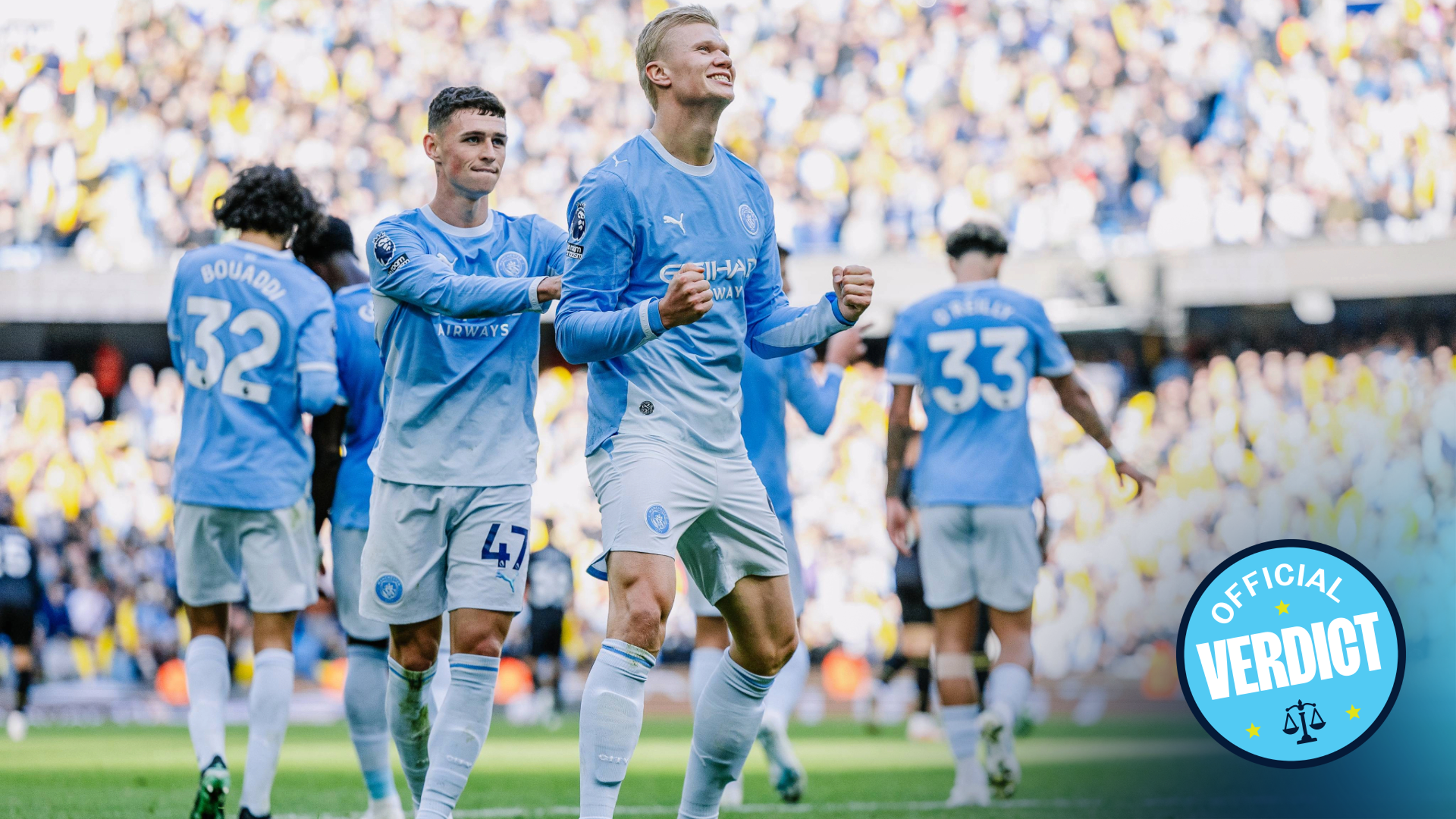 A group of Manchester City players celebrating a goal on the field. One player is prominently fist-pumping while others in light blue kits are in the background.