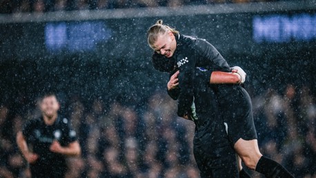 Two football players wearing black jerseys hug each other during a game played in the rain, with another player visible in the background.