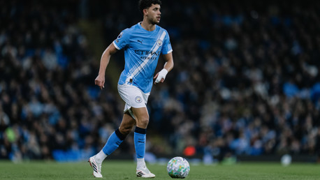 A Manchester City player in the team's kit is on a soccer field with a blurred face, holding a football.