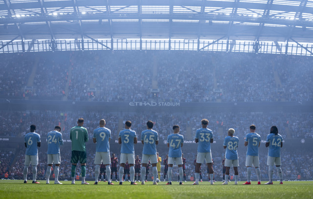 Manchester City team lined up in front of a packed Etihad Stadium. Players are wearing light blue jerseys, facing the crowd.