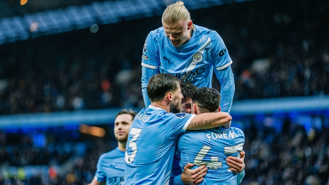 Manchester City players celebrating a goal in their iconic blue jerseys. Phil Foden is visibly seen with Ruben Dias and another player, while a blurred face of a player on top of the group is present.