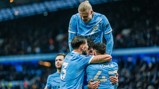 Manchester City players celebrating a goal in their iconic blue jerseys. Phil Foden is visibly seen with Ruben Dias and another player, while a blurred face of a player on top of the group is present.