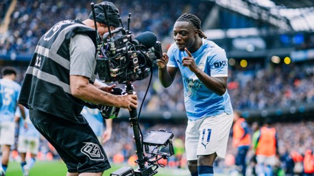 A Manchester City player is interacting with a camera operator during a match at the Etihad Stadium. The player is wearing Manchester City's home kit.