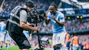 A Manchester City player is interacting with a camera operator during a match at the Etihad Stadium. The player is wearing Manchester City's home kit.