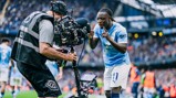 A Manchester City player is interacting with a camera operator during a match at the Etihad Stadium. The player is wearing Manchester City's home kit.