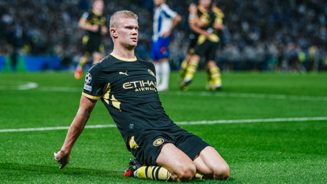 A Manchester City player in a black and yellow jersey, celebrating by sliding on the pitch. The jersey features Etihad Airways as the sponsor.