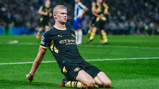 A Manchester City player in a black and yellow jersey, celebrating by sliding on the pitch. The jersey features Etihad Airways as the sponsor.