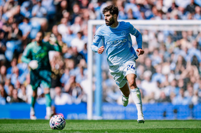 A soccer player in a Manchester City uniform running on the field during a match. The background shows the stadium with spectators.