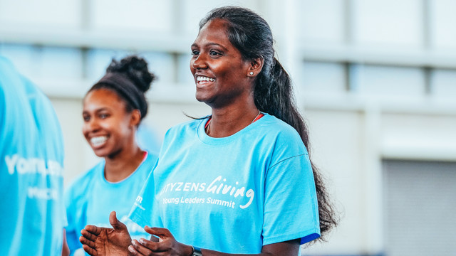 A person in a blue 'Cityzens Giving Young Leaders Summit' t-shirt claps during an event. The background suggests an outdoor gathering.