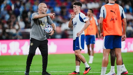 A soccer coach giving instructions to a player holding a ball during a training session. Other players are in the background.