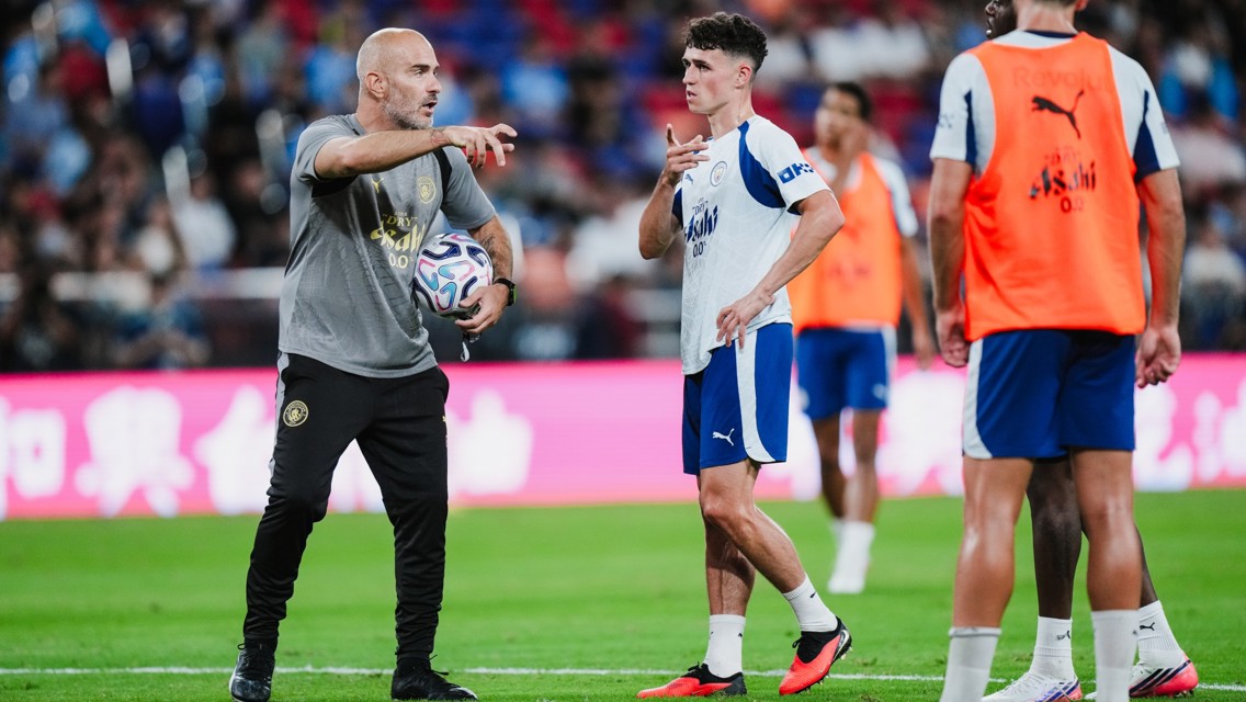 A soccer coach giving instructions to a player holding a ball during a training session. Other players are in the background.